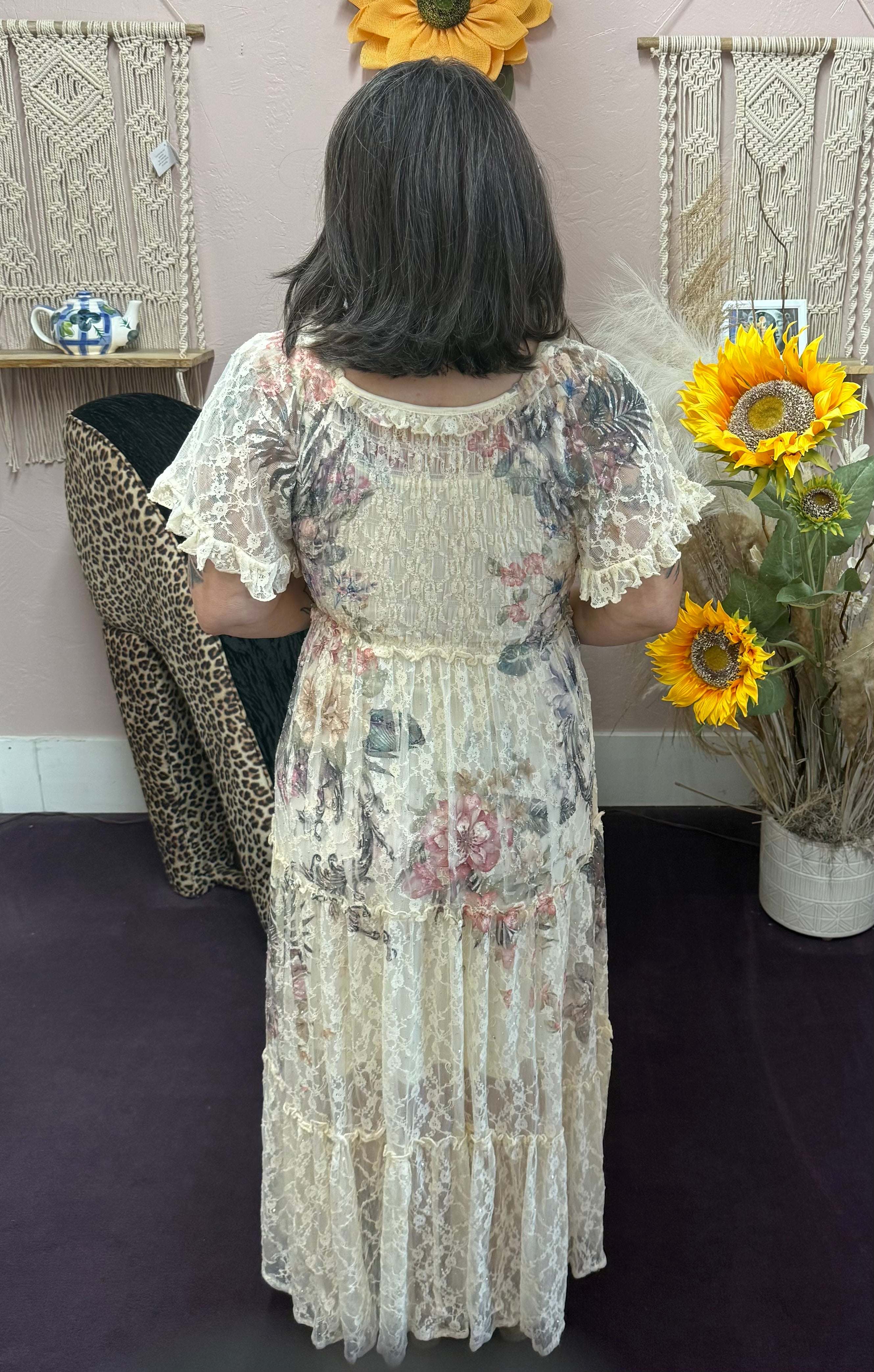 Person wearing a floral dress holding sunflowers in a room with decorative wall hangings.
