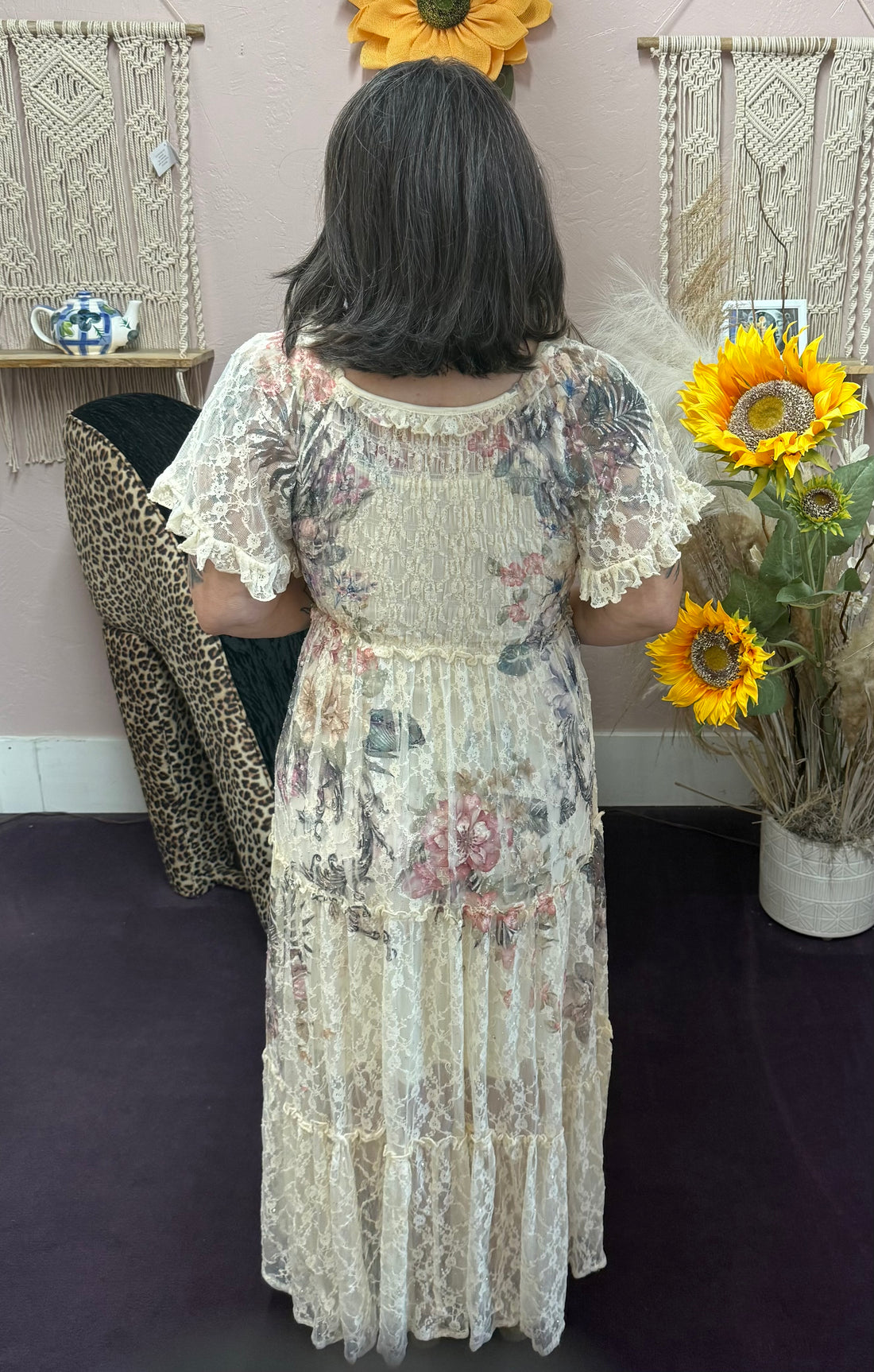 Person wearing a floral dress holding sunflowers in a room with decorative wall hangings.