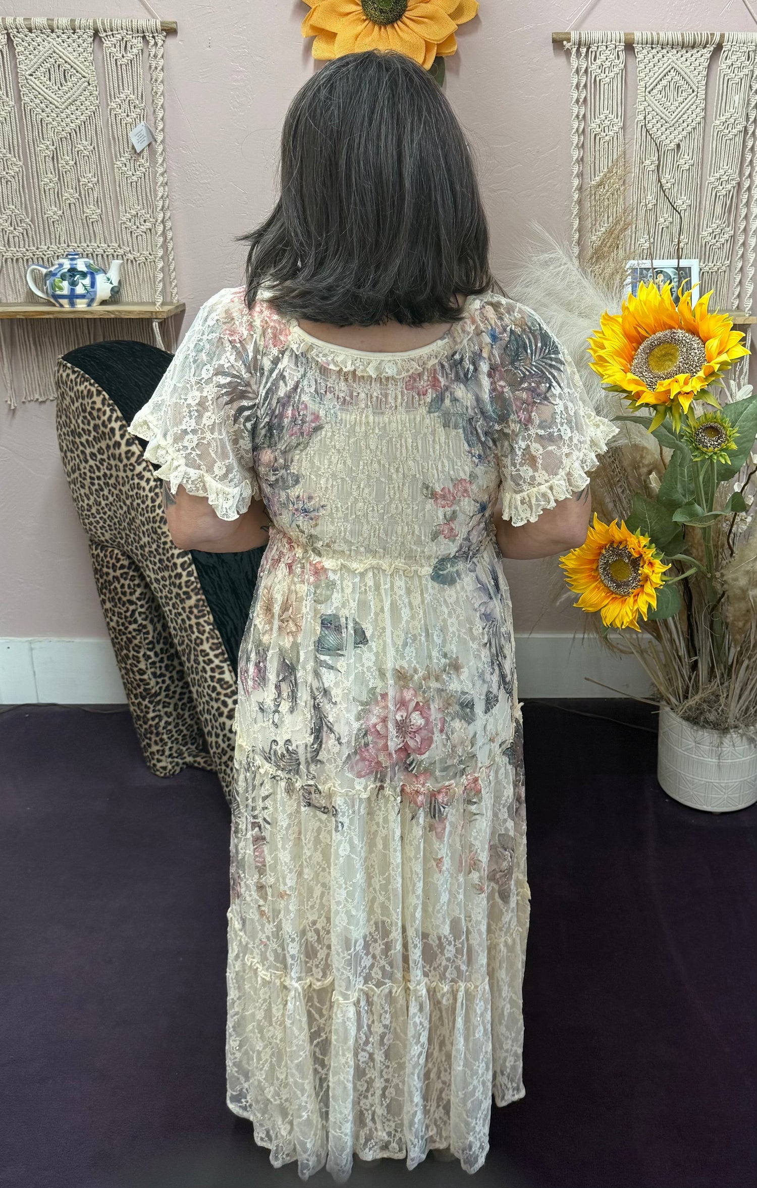 Person wearing a floral dress holding sunflowers in a room with decorative wall hangings.