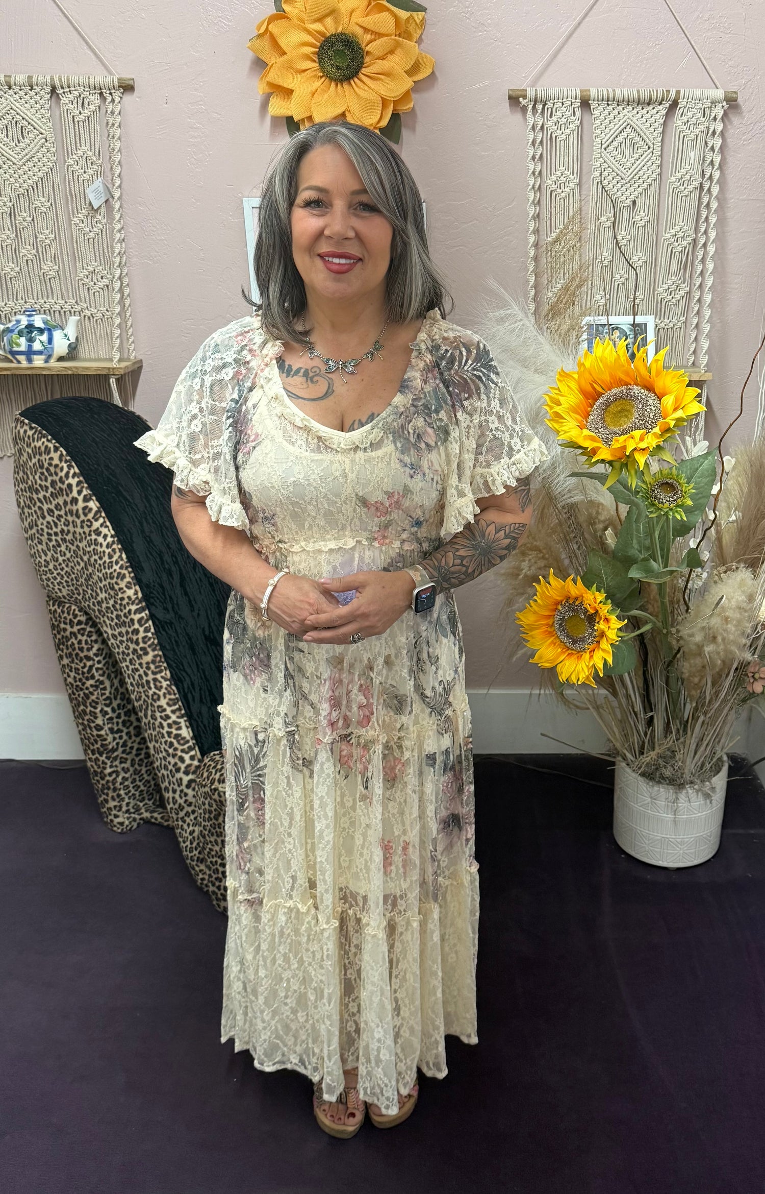 Woman in a floral dress standing in front of decorative wall hangings with sunflowers.